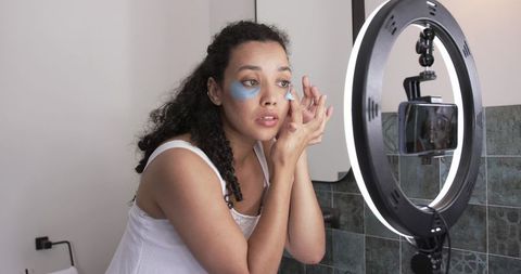 African american woman applying under-eye patches filming skincare routine with ring light