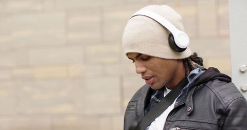 Young man leaning against column listening to music with headphones in urban streetwear