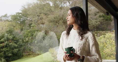 Young woman standing by glass window holding mug, gazing contemplatively at garden view