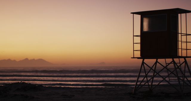 Lifeguard tower standing alone on a sandy beach during sunset with an orange and pink sky, mountains barely visible in the background. Ideal for use in travel brochures, beach-themed decor, relaxation and wellness articles, or summer vacation promotions.