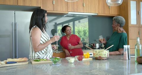 African american women sharing wine and prepping salad around modern kitchen island