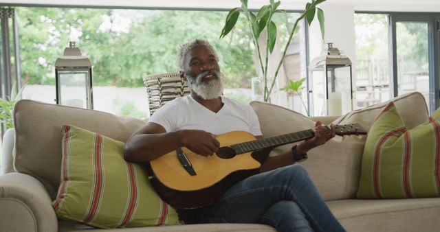 A senior man with a long white beard plays an acoustic guitar while sitting on a cozy couch in a bright living room. The relaxed setting includes green pillows, indoor plants, and large windows letting in natural light. This image is perfect for representing leisure at home, musical hobbies, or promoting a serene lifestyle for older adults.