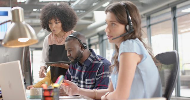 Team of diverse coworkers collaborating in a modern office, two wearing headsets while managing customer service tasks, third person reviewing information on tablet. Ideal for use in corporate presentations, customer support services advertising, and teamwork-oriented settings.