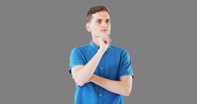 Young man standing with hand on chin, wearing a blue shirt, against a gray background. Image is useful for concepts like contemplation, thinking, decision-making, and introspection. Perfect for websites, blogs, or advertising related to inspiration, focus, and problem-solving.