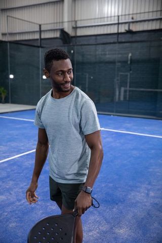 African american man playing padel on indoor blue court wearing smartwatch