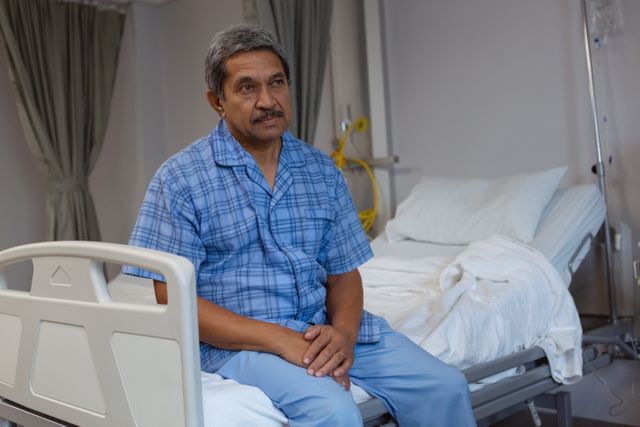 Front view of mature male patient sitting on medical bed and looking away in medical ward at hospital