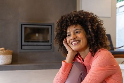 African american woman smiling and relaxing by modern fireplace in sunlit living room