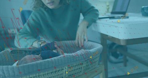 Working mother monitoring newborn in bassinet near laptop with real-time health data overlay