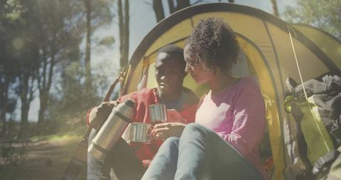 Sunlit couple camping sharing coffee by yellow tent, cozy intimate morning in forest