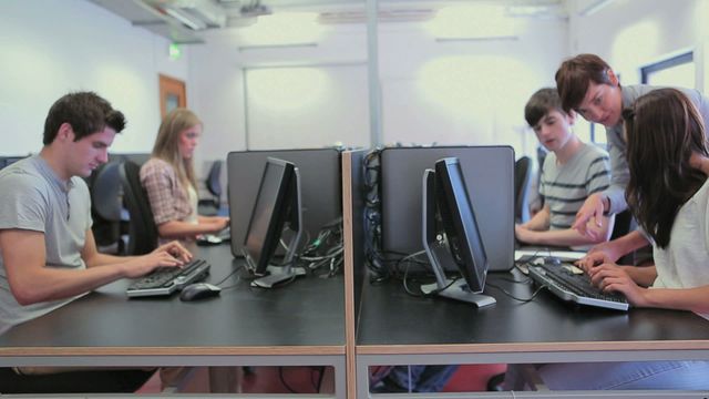 Students and teacher engaged in computer-based learning in a modern classroom. Young adults receiving guidance in technology education, perfect for content on academic environments, modern education systems, teamwork, and tech-based learning. Suitable for promoting educational institutions, classroom interactions, and collaborative learning tools.