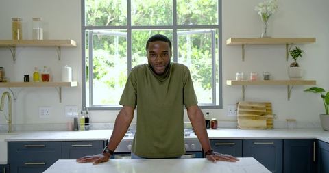 Young man leaning on marble island in bright modern kitchen with window light and greenery