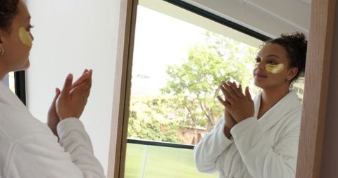 African american woman applying gold under-eye patches wearing white robe at mirror
