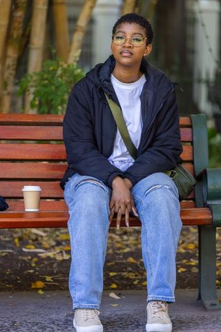 African american woman sitting on bench wearing glasses, ripped jeans and puffer jacket