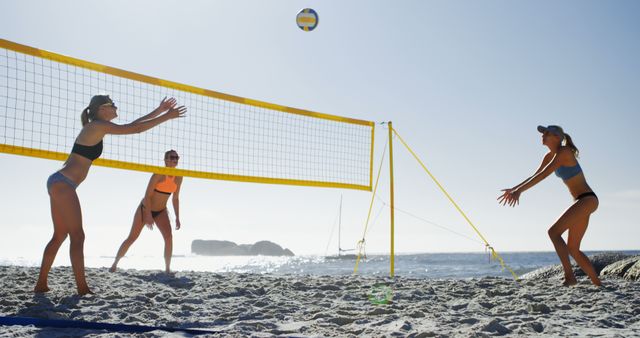 Three women in swimwear engaged in a lively game of beach volleyball on a sunny day. They are positioned around the net with focus on the ball, showcasing teamwork and athleticism. Perfect for topics on outdoor sports, summer activities, fitness, and beach vacations to create energetic and spirited content.