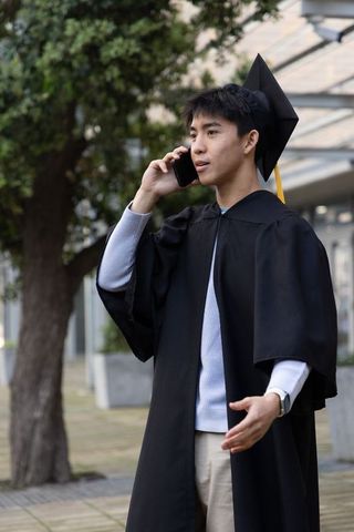 Asian graduate wearing mortarboard and gown talking on phone on campus path
