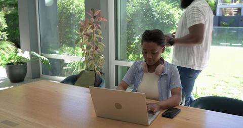 Young woman typing on laptop at sunlit table with greenery, man checking watch