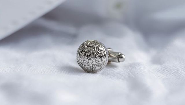 Engraved silver cufflink on white shirt fabric showing ornate paisley detail and luxury
