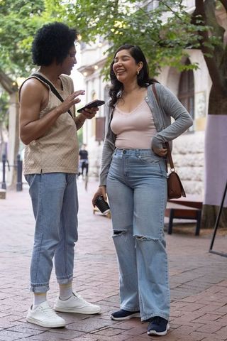 Couple sharing laugh while checking smartphone and holding coffee on urban brick street