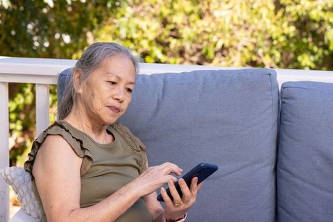 Senior asian woman relaxing on sunlit patio sofa using smartphone for connection