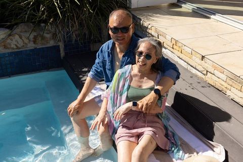 Senior asian couple relaxing by pool dipping feet, laughing on patio deck in summer