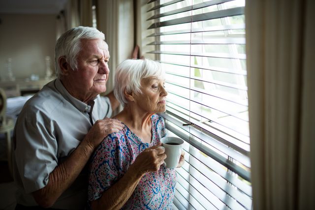 Senior couple standing together, looking out window, conveying themes of togetherness, contemplation, and peaceful retirement. Suitable for use in articles or advertisements related to aging, retirement living, senior care, and family bonding.