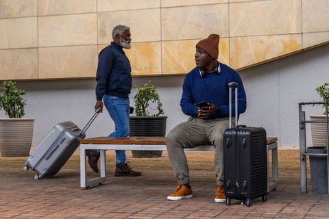 Urban travelers resting on bench with suitcases, headphones and smartphone in transit plaza courtyar