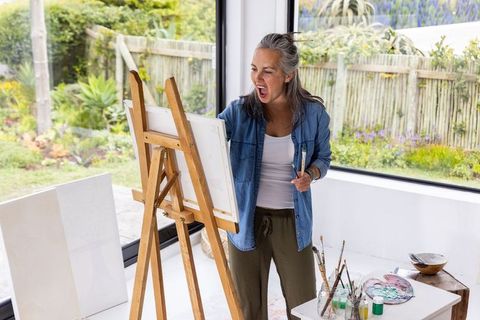 Senior woman painting at easel in sunlit home studio with garden view and palette
