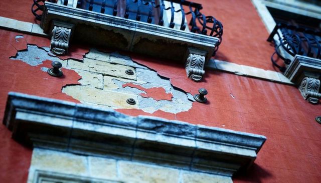 Peeling red stucco revealing yellow underlayer with weathered cornices and iron balconies