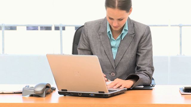 Businesswoman using a laptop in a corporate office space. She is wearing professional business attire. Ideal for themes involving business, technology, productivity, and professional work environments.