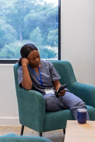 African nurse taking break in teal armchair checking smartphone in hospital lounge