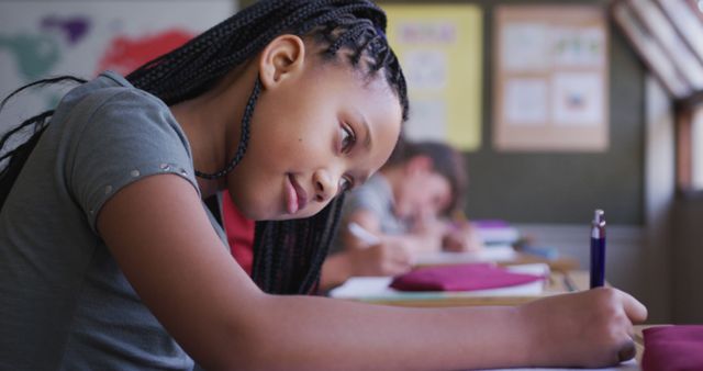 African girl sitting at desk writing in notebook with other students in background. Captures focused academic effort and engagement, ideal for educational materials, school brochures, online learning platforms, and articles on childhood education.