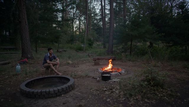 Sitting camper tending campfire at pine forest campsite during twilight with cooking pot