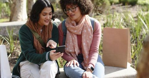 Two young women sharing smartphone while sitting on park bench, casual outdoor friends
