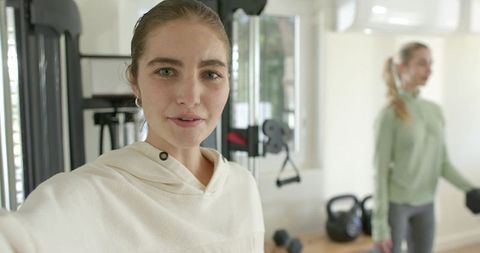 Young women taking selfie in bright home gym with kettlebells, dumbbells, and cable machine