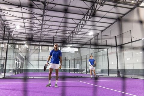 Male teammates playing padel doubles on purple indoor court with glass walls and net