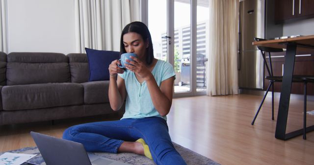 Biracial gender fluid person sitting on floor drinking coffee and using laptop at home - Download Free Stock Photos Pikwizard.com