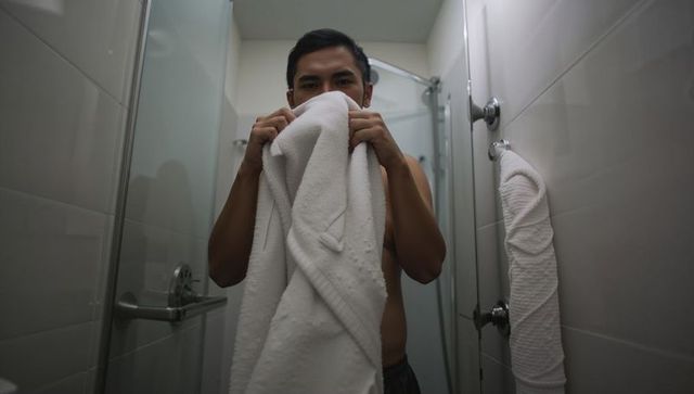 Young asian man drying face with white towel in minimalist glass shower, neutral bathroom