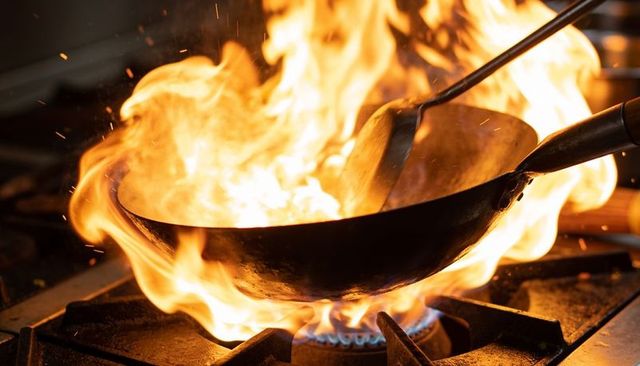 Wok flaming over gas burner with high heat flame, spatula tossing ingredients