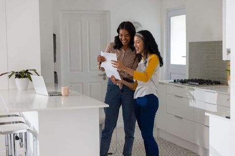 Diverse mother and teenage daughter sharing documents and smiling in modern kitchen