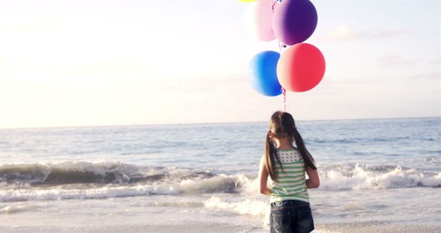 Girl holding colorful balloons at the edge of the beach with ocean waves in the background. Good for themes of childhood, summer vacations, freedom, and outdoor fun.