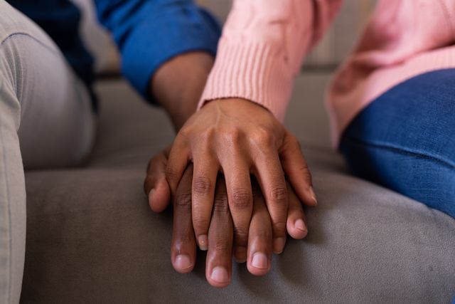 This image shows a close-up of a biracial couple holding hands while sitting on a couch at home. It can be used to represent themes of love, unity, and inclusivity in relationships. Ideal for articles, blogs, and advertisements focusing on interracial relationships, domestic life, and emotional support.
