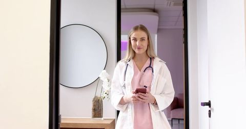 Clinician standing in clinic doorway holding smartphone wearing pink scrubs and lab coat