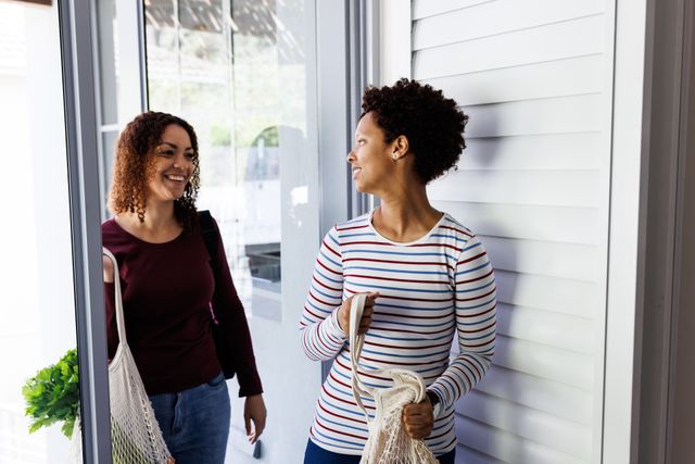 Two women, one with curly hair and the other with short hair, are arriving home with reusable shopping bags. They are smiling and talking in the doorway, suggesting a close relationship and shared domestic life. This image can be used to depict themes of togetherness, domestic life, and eco-friendly shopping. Ideal for use in lifestyle blogs, advertisements promoting reusable bags, or content celebrating LGBTQ+ relationships.