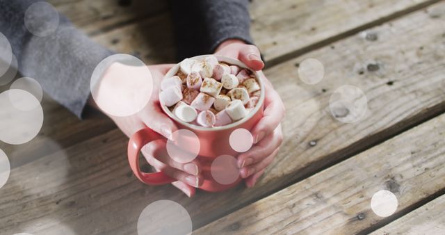 Warm hands holding a red mug filled with hot cocoa and marshmallows on a rustic wooden table. The warm glow of light bokeh creates a cozy atmosphere. Perfect for use in holiday greeting cards, winter promotions, cozy home decor themes, and social media content celebrating the winter season.