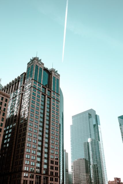 Image capturing towering modern skyscrapers against a clear sky with a visible contrail. It can be used for themes related to urban development, corporate office locations, real estate, and metropolitan skylines. Perfect for illustrating business growth, architectural design, and skyline studies.