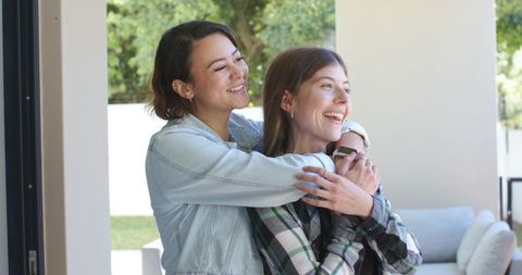 Diverse female friends hugging and laughing on sunny patio, denim jacket and smartwatch