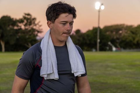 Young athlete standing on grassy field with towel at dusk under stadium floodlights