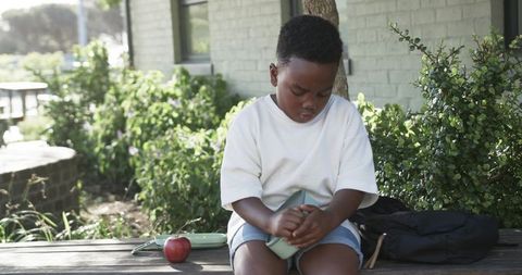 School child opening lunchbox on bench during outdoor lunch break with backpack and apple