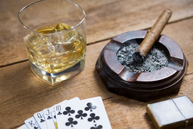 This image captures a scene of relaxation and leisure with a glass of whisky, a lit cigar in an ashtray, and playing cards on a wooden table. Ideal for use in articles or advertisements related to luxury lifestyle, relaxation, gambling, or social gatherings. It can also be used in blogs or websites focusing on whisky, cigars, or card games.