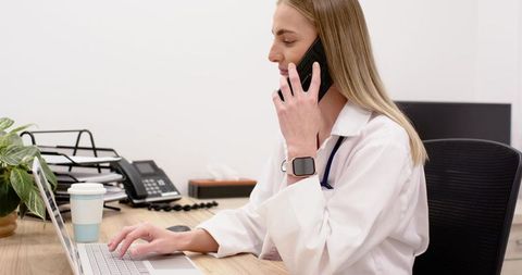 Female physician talking on phone and typing on laptop in modern clinic office